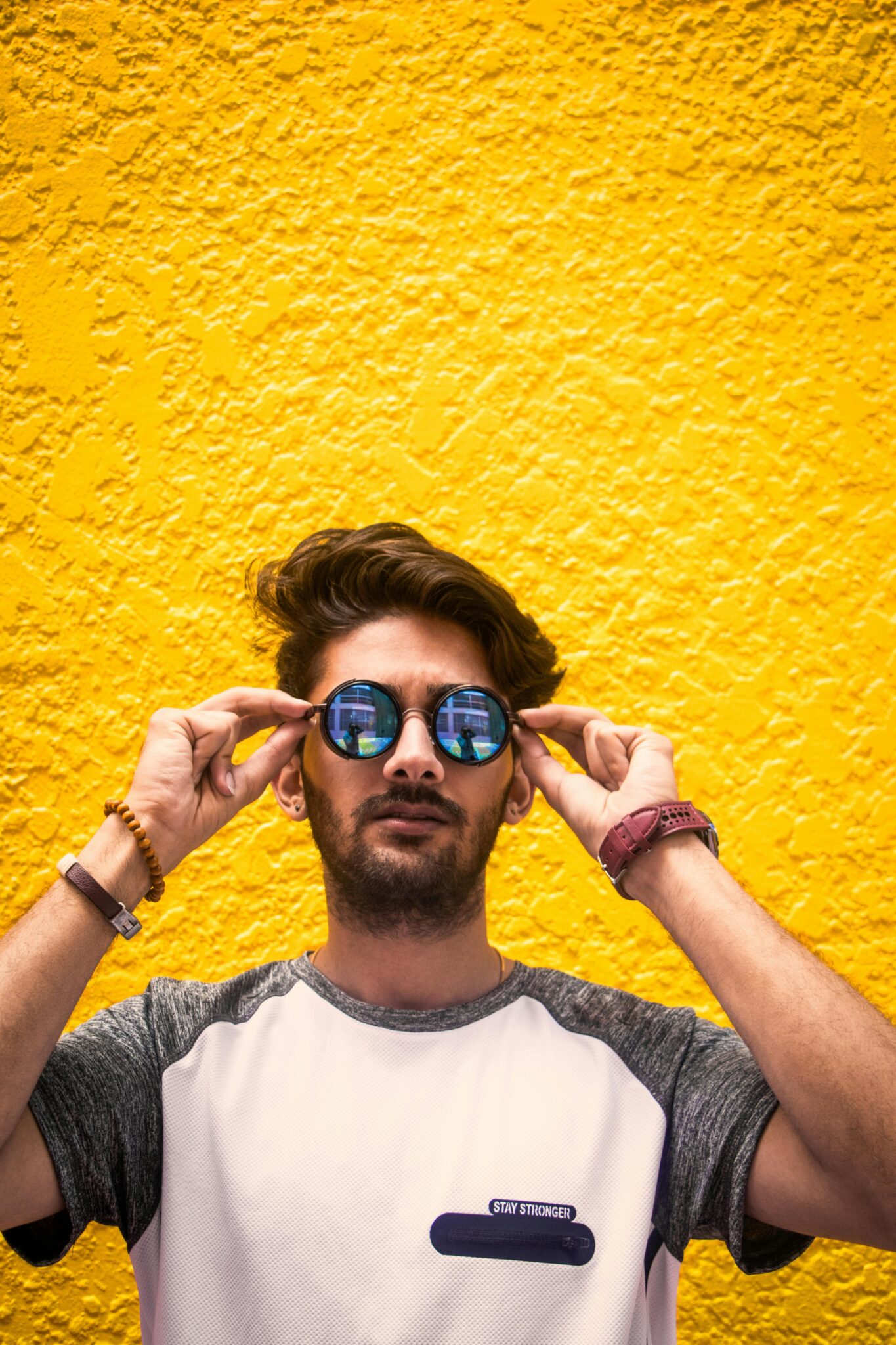 Close-up of a young, bearded man wearing round reflective sunglasses against a vibrant yellow wall.