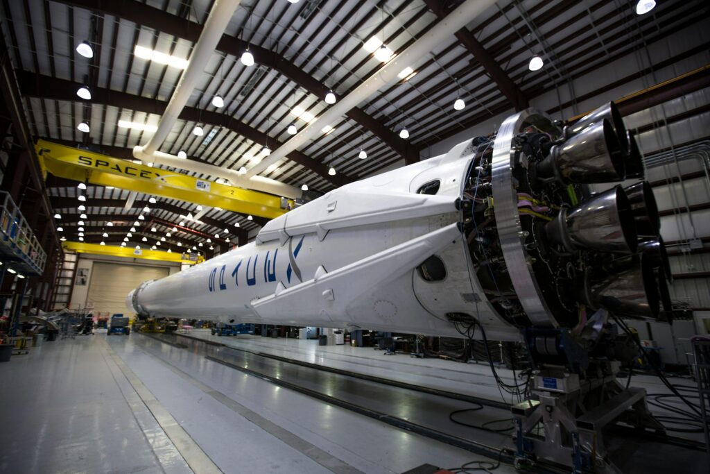 A horizontally positioned SpaceX rocket inside a large industrial facility, showing its engines and assembly equipment in a well-lit hangar.