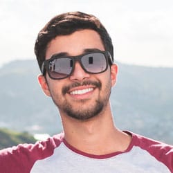 Smiling man with short, dark hair wearing sunglasses and a maroon-gray T-shirt against a scenic backdrop.